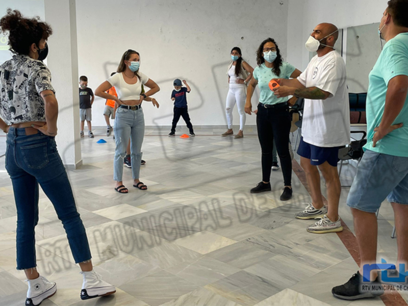 Un grupo de personas, incluyendo niños y adultos, participa en una actividad al aire libre con un fondo de edificios. La imagen muestra a los participantes en una zona abierta, posiblemente un parque o espacio público. La iluminación sugiere que es una tarde soleada, y la presencia de sombrillas en el suelo indica que se está protegiendo del sol. La gente parece estar disfrutando de la actividad, con algunos sujetos mirando hacia el frente y otros alrededor.