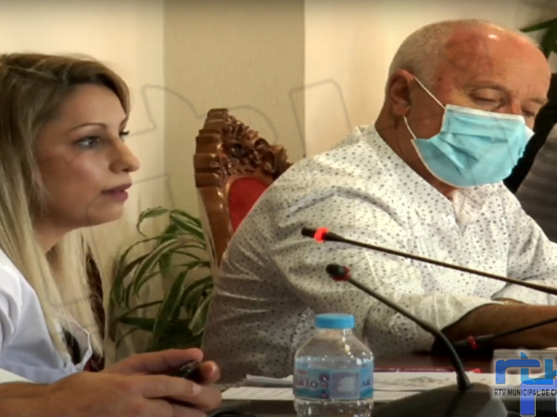 Dos personas con máscaras faciales, una mujer y un hombre, están sentados en una sala de conferencias. Hay micrófonos en la mesa y un botellín con agua. La imagen parece ser de una reunión o conferencia formal, posiblemente relacionada con la salud pública.