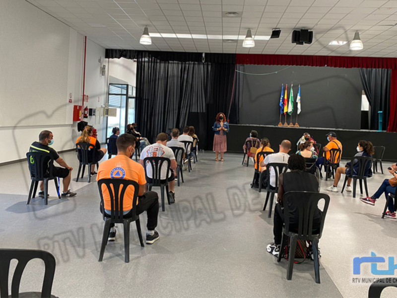 Un grupo de personas sentadas en sillas negras, mirando a una persona que está hablando frente al escenario. La sala tiene un techo con luces y una cortina negra en el fondo.