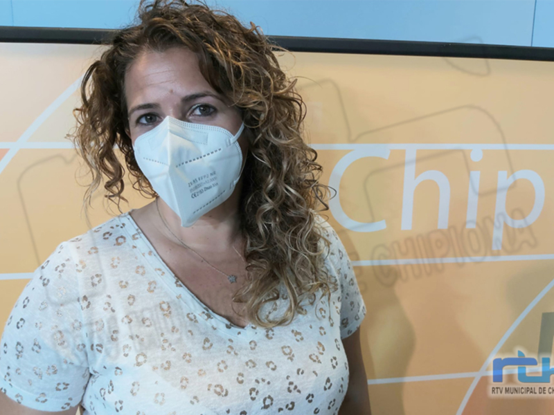 A una mujer con cabello rizado y una máscara facial blanca, posiblemente en un entorno de trabajo o salud pública. La pared al fondo tiene una publicidad con el nombre "Chipiona" y la imagen de un teléfono móvil.