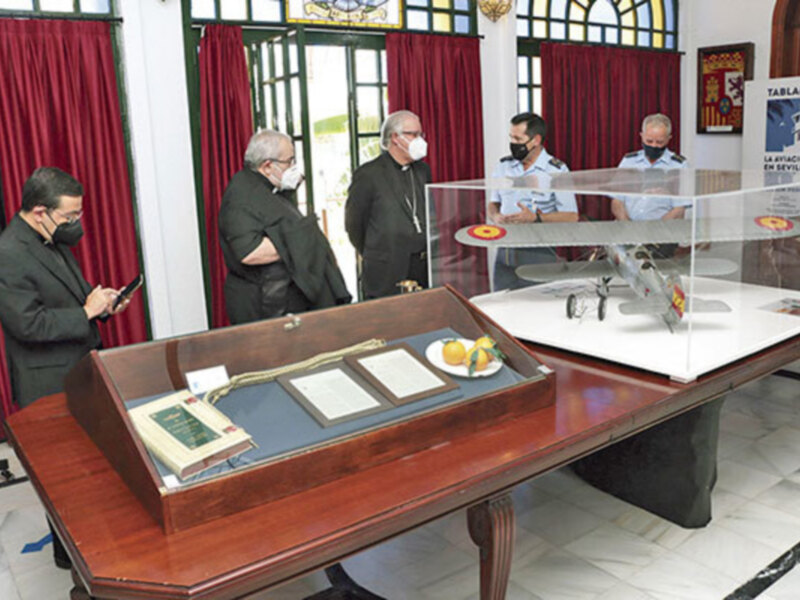 Exposición de documentos y modelos aéreos en un espacio con decoración clásica y personas observando.
