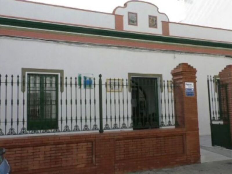 Fachada de una casa blanca con detalles verdes y rojos, puerta principal con barrotes metálicos. Detalle de la calle y un coche parado a la derecha.
