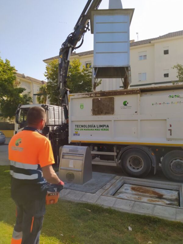 Un trabajador en uniforme naranja y blanco está operando un camión de basura con una grúa, mientras recoge residuos en una calle. La imagen muestra la tecnología limpia para un manejo más verde, enfatizando el uso de tecnologías modernas en la gestión de residuos.