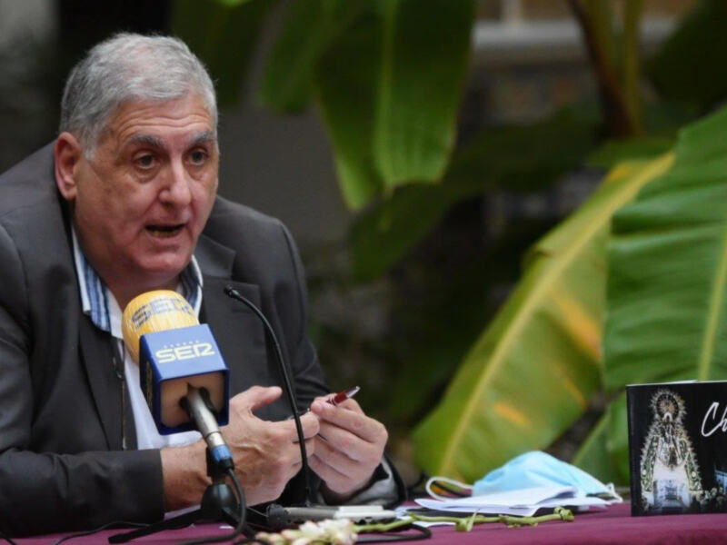 Hombre hablando en rueda de prensa con micrófono y libro de 'Regla de Chipiona' en la mesa.