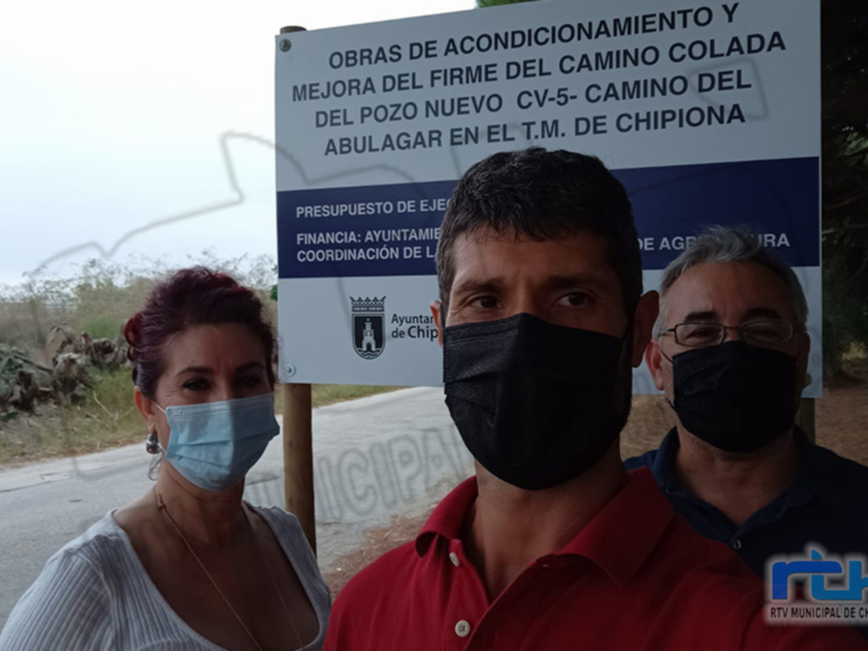 Se observa a tres personas, dos de ellas con mascarillas faciales, posando frente a un cartel informativo sobre obras de acondicionamiento y mejora del firme en el camino Colada, ubicado en Chipiona. El cartel indica que se trata de una obra financiada por la Ayuntamiento de Chipiona, con presupuesto ejercido y coordinación de la Coordinación de Agricultura.