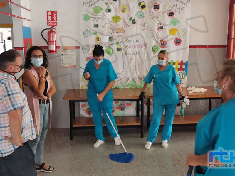 En una escuela, dos personas en uniforme azul limpiando el piso con un paño de microfibra. A la izquierda, dos personas observan la limpieza. En el fondo, un mural educativo con dibujos y texto en español.