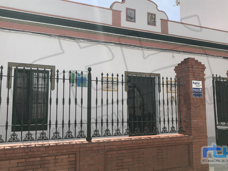 Fachada de una casa blanca con ventanas y puertas protegidas por un enrejado metálico. La casa tiene detalles de ladrillo rojo y una inscripción en la puerta.