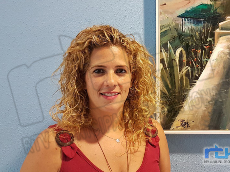 A una mujer con cabello rizado y rubio, sonriendo frente a un fondo azul. A su lado se encuentra una pintura que representa una casa en el campo, con un cielo rojizo. La imagen parece estar tomada en un evento o presentación, como lo indica el logo visible en la esquina inferior derecha.
