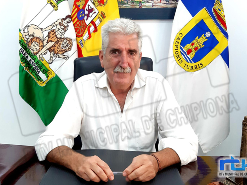 Un hombre con una camisa blanca está sentado en un escritorio, frente a varias banderas y documentos. La imagen parece estar relacionada con el gobierno o la administración municipal de Chipiona, según se puede leer en los textos y símbolos visibles.