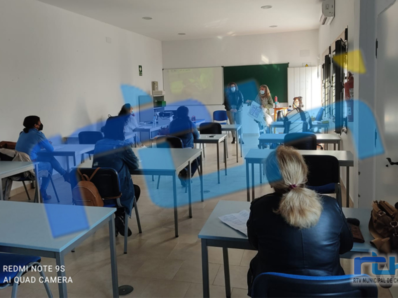 Salón de clase con estudiantes sentados en mesas y sillas, paredes blancas, pizarra verde, ventana con cortinas.