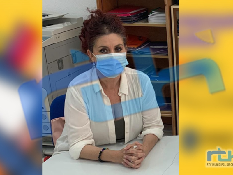 A una persona sentada en un escritorio, con una camisa blanca y una bufanda azul. La persona lleva una mascarilla facial. En el fondo, se pueden ver estanterías con libros y otros objetos de oficina. La imagen está borrosa en los bordes, lo que sugiere una captura de movimiento o un efecto de desenfoque.