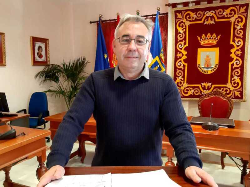Un hombre con una camisa azul y gafas está sentado en un escritorio de madera. A su lado, hay una bandera y un escudo heráldico en el fondo. La imagen parece estar tomada en una oficina o lugar de trabajo formal.