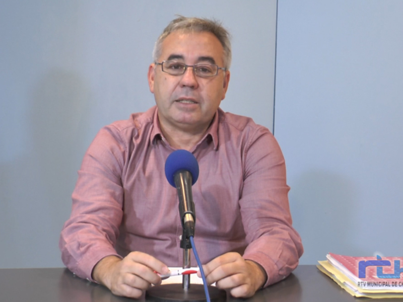 Un hombre con cabello gris y gafas de montura metálica habla en un micrófono azul. Está sentado frente a una mesa con documentos y un cuaderno en el lado derecho. El fondo es un muro azul claro con una sombra de la persona en el suelo.