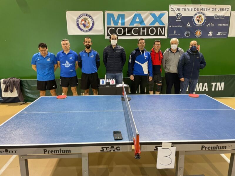 Un equipo de tenis de mesa posa frente a una mesa azul en un club deportivo, con banners y logotipos de patrocinadores en el fondo.