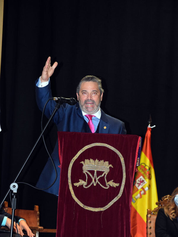 Un hombre en traje y corbata roja se dirige a un público, sosteniendo una bandera española en el fondo.