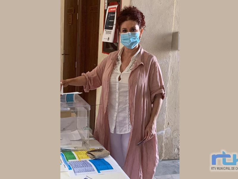 Una mujer votando en un colegio electoral, con una máscara facial y vestida con un abrigo rosa. La elección se lleva a cabo en Chipiona, con un marcador de votación visible en el fondo.