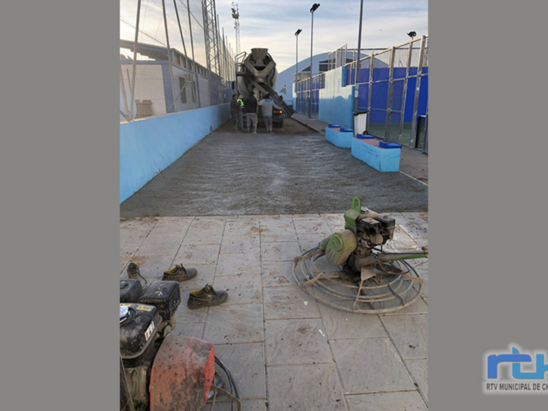 Una máquina de hormigón se encuentra en el centro de la imagen, con un muro azul y una estructura metálica al fondo. En el suelo, hay varios cascos de construcción y una máquina de hormigón parcialmente visible. La escena parece estar en un área industrial o construcción, con una pista de hormigón y un cielo despejado.