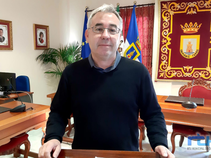 Un hombre con una camiseta negra y gafas de prescripción se encuentra en un despacho. En el fondo, hay una bandera europea y una bandera nacional, junto con un escudo de armas. La oficina está decorada con una planta y dos cuadros en la pared, uno de los cuales muestra a un hombre. En el escritorio hay una computadora y otros objetos de oficina.