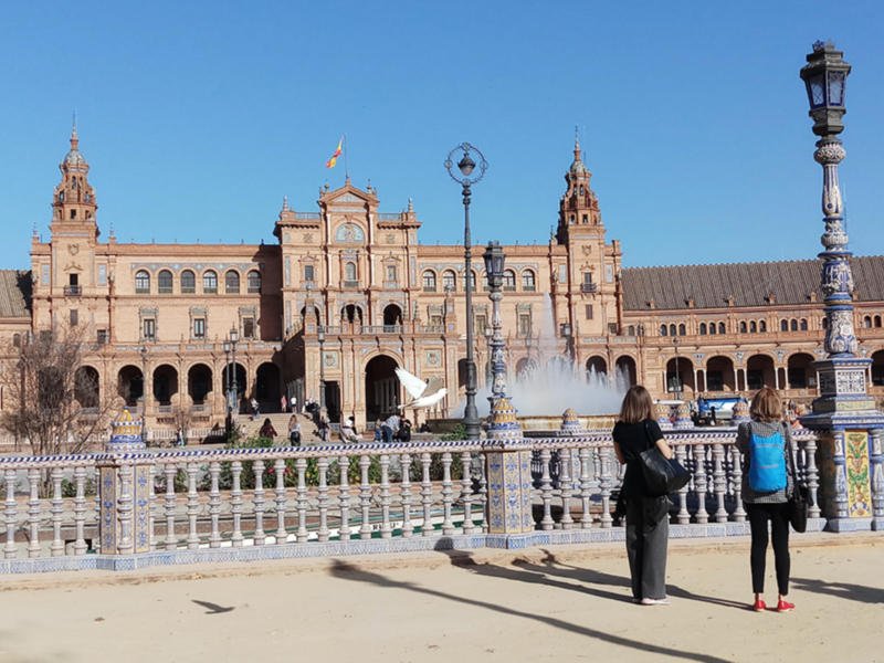 Plaza de España en Sevilla, España, con palomas volando y dos personas en el centro del parque.