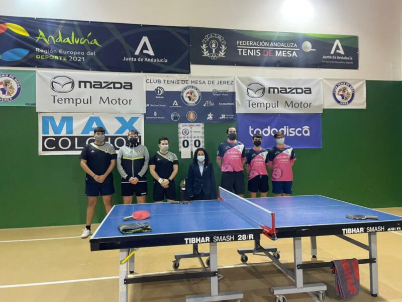 Equipo de tenis de mesa posando en la cancha con banners de sponsors y la bandera de Andalucía en el fondo.