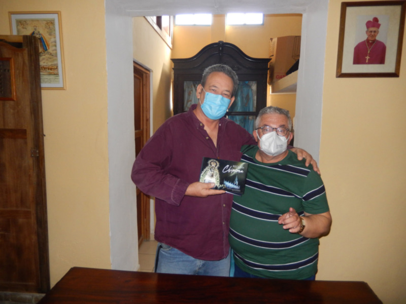 Dos hombres con mascarillas posan juntos, uno sostiene un libro titulado 'Recuerdos de Chipiona'.