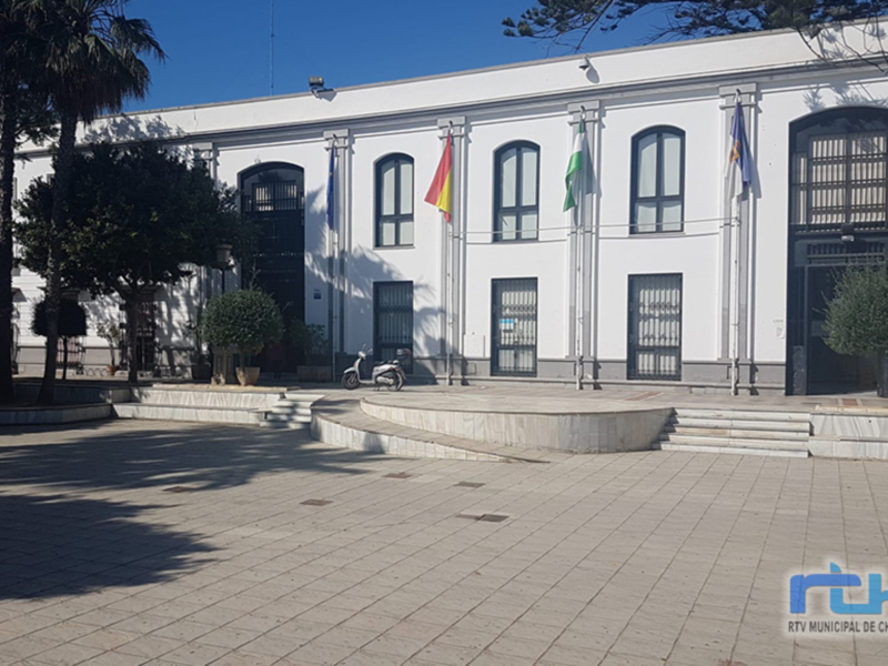 Edificio blanco con ventanas y banderas en la entrada, frente a un parque con árboles.