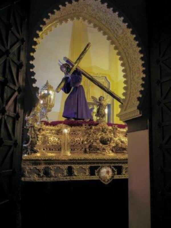 Un altar elaborado con detalles dorados y una figura vestida de morado sosteniendo una cruz. La decoración es sofisticada, con un marco de madera y detalles en oro que reflejan lujo y esplendor. La iluminación es suave, enfocándose en el altar centralizado.