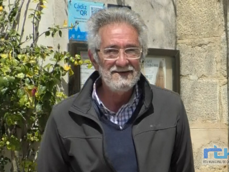 Un hombre con barba y gafas, vestido con una chaqueta de color oscuro, frente a un edificio con una placa que dice "Cádiz en QR". La imagen muestra detalles de su rostro y parte del edificio.
