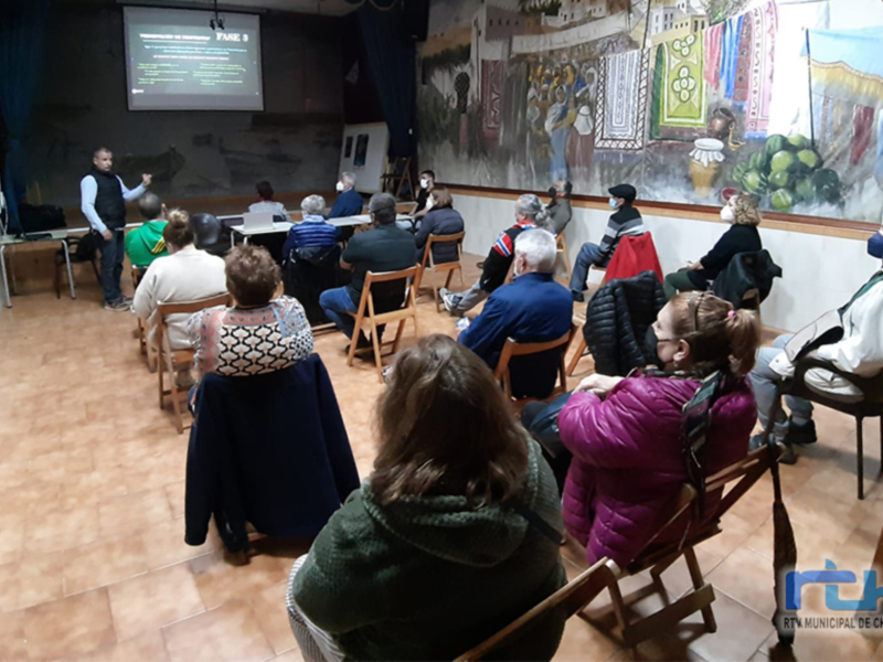 Una sala de conferencias con una pantalla proyectando 'FASE 3' mientras una persona habla a una audiencia de personas sentadas en sillas. La sala está decorada con murales y carteles en las paredes.
