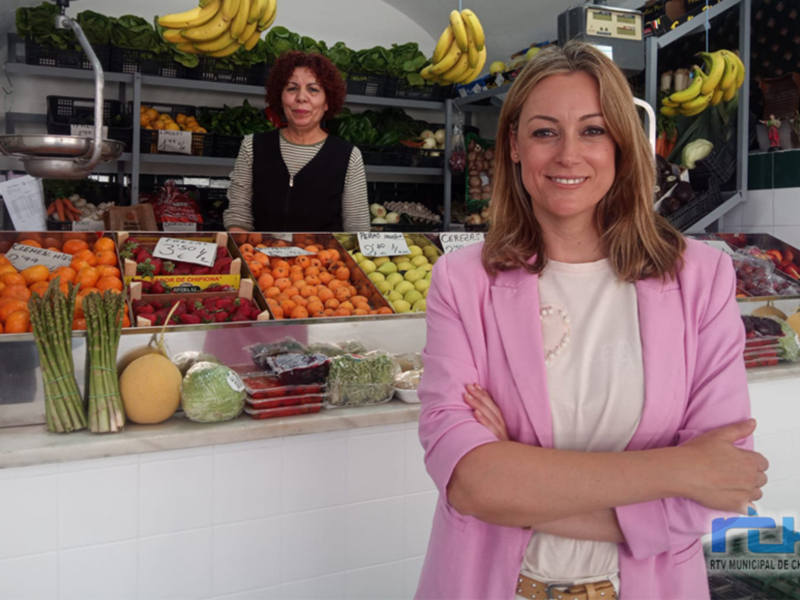 En un mercado de frutas y verduras, una mujer con cabello largo y una blusa rosa sonríe mientras las manos cruzadas. A su lado, una mujer con cabello rojo y una blusa negra observa el ambiente. En el fondo, se pueden ver frutas como naranjas, plátanos y verduras frescas. La imagen destaca la variedad de productos disponibles en el mercado.