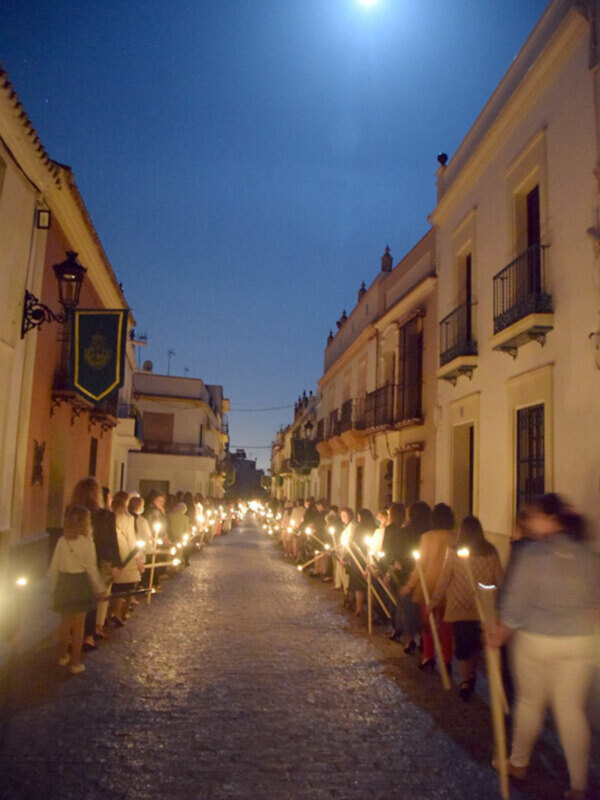 Una imagen nocturna de una calle adoquinada con gente formando una fila, cada uno con un farol en la mano. El cielo está claro y brillante con una luna grande. Las casas a ambos lados de la calle tienen balcones y ventanas con detalles arquitectónicos. La atmósfera es tranquila y festiva, probablemente en una celebración o procesión nocturna.
