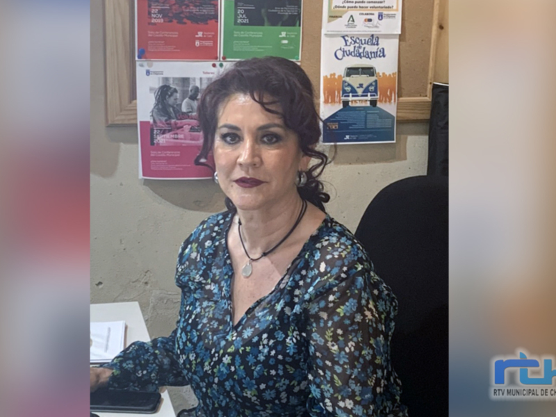 A una mujer sentada en un escritorio, con una computadora y varios carteles de eventos en la pared detrás de ella. La mujer lleva una camisa con estampado floral y un collar, y parece estar en un entorno profesional o de trabajo.