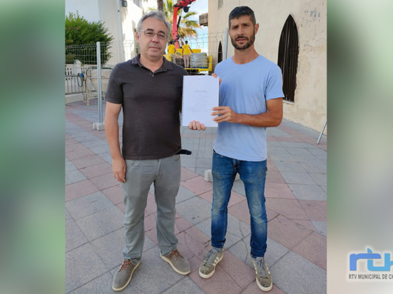 Dos hombres posan con un papel en la mano, frente a una estructura de hormigón. El fondo muestra una calle con un poste de luz y edificios de estilo colonial. La imagen parece ser de Chipiona, Andalucía, España.
