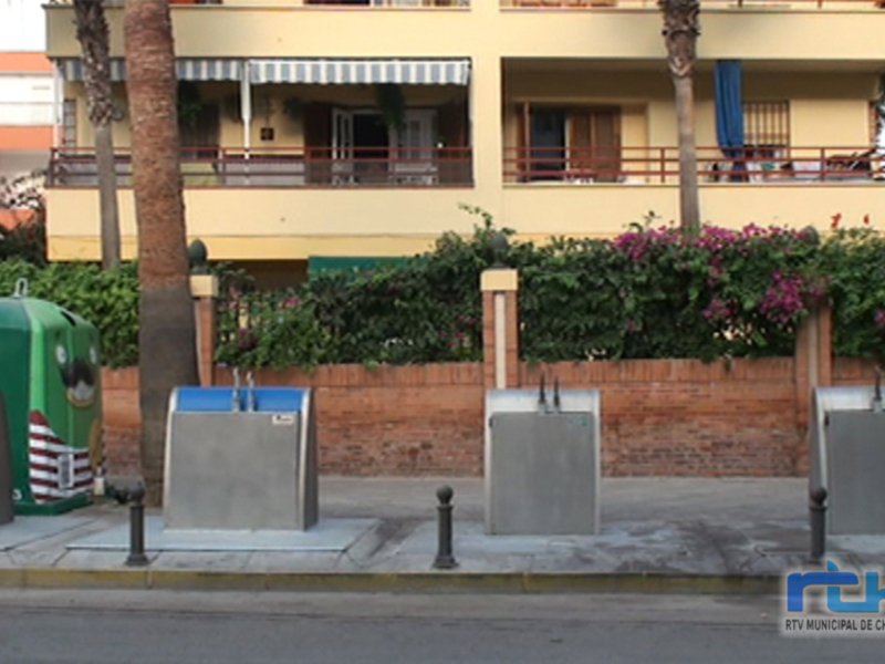Edificio con balcones y toldos, jardín con flores, contenedores de basura en la calle.