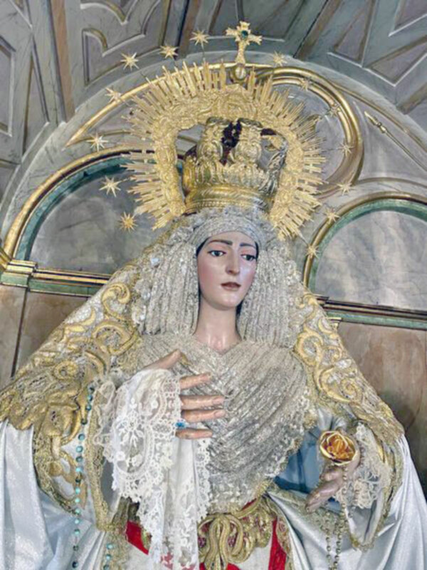 Una estatua de la Virgen María, vestida con un manto blanco y dorado, sosteniendo una rosa. La estatua está coronada con un halo de estrellas y una corona dorada, rodeada por una arquitectura de estilo barroco con detalles dorados y azules. La escena transmite una sensación de solemnidad y devoción religiosa.