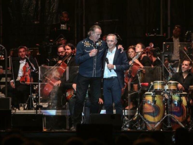 Artistas en escenario con orquesta de fondo durante un concierto.