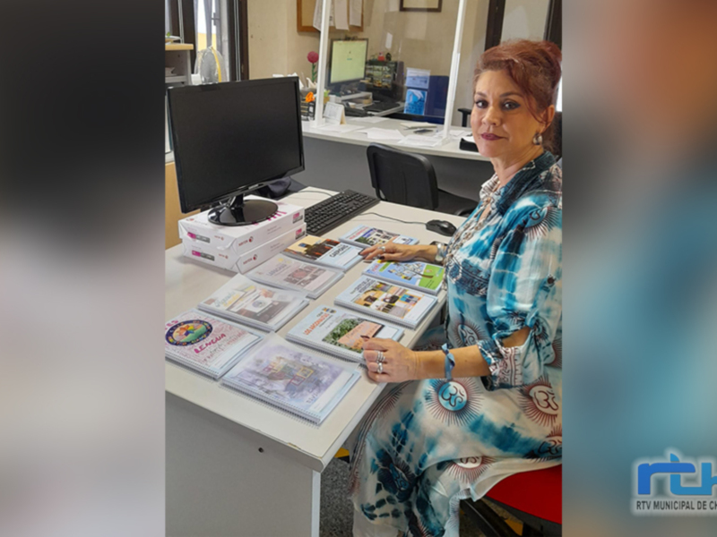 Se muestra a una mujer sentada en un escritorio, frente a un computador y varios documentos. La escena parece ser una oficina o lugar de trabajo, con elementos como un monitor, teclado y estantes en el fondo. La mujer está sonriendo y parece estar trabajando o presentando algo.