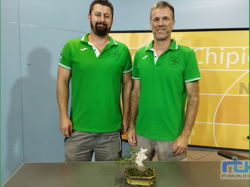 Dos hombres vestidos con camisetas verdes de la misma marca posan frente a una pared gráfica. La imagen incluye un bonsái en el suelo y la marca "RTV Municipal de Chipiona" en la esquina inferior derecha.