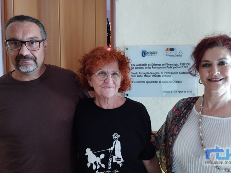 Una imagen de un evento donde una persona con cabello rojo y dos personas más son reconocidas por sus logros en la Asociación de Enfermos de Fibromialgia. La foto está firmada por la televisión municipal de Chipiona.