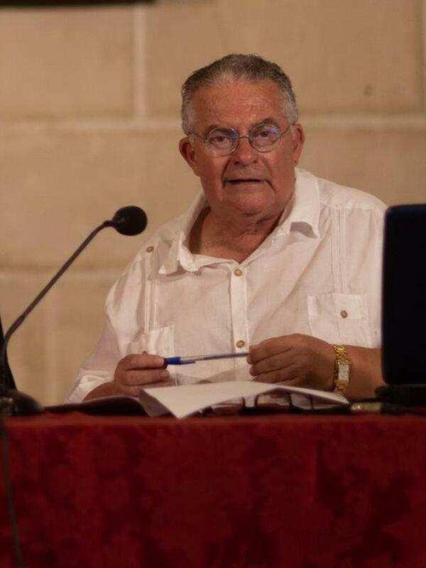 Un hombre en camisa blanca está leyendo un libro mientras sostiene una pluma. Está sentado frente a un fondo beige y tiene un portátil en su regazo.