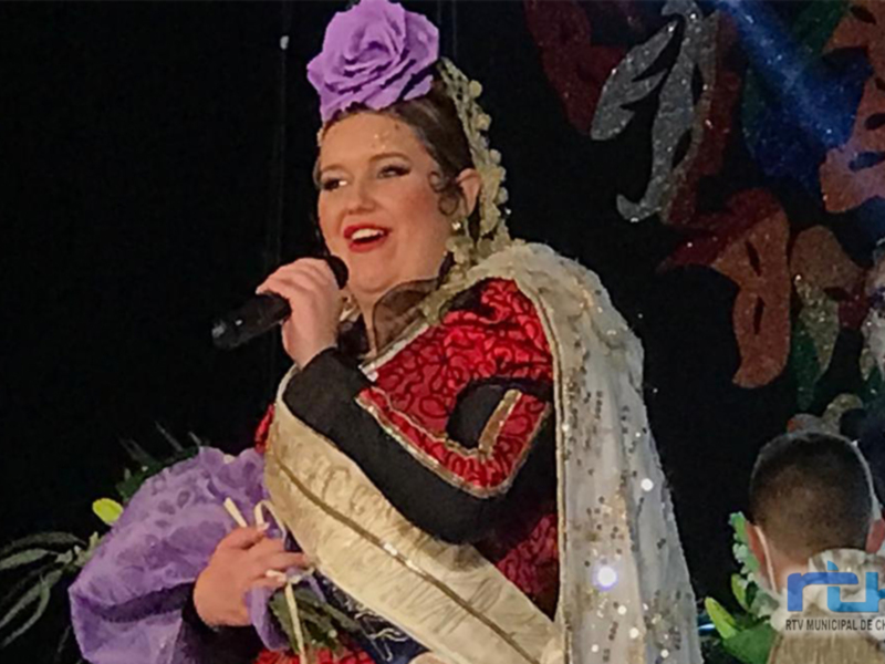 Una mujer con un vestido tradicional de color rojo y blanco, llevando una corona de flores en la cabeza. Está cantando con un micrófono en mano, y hay una decoración festiva al fondo.