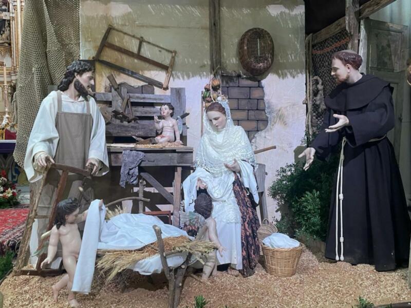 Escena navideña con figuras de madera representando a la Virgen María, el Niño Jesús y un santo franciscano. En el fondo, una estatua de una iglesia y una escena con un niño en un balcón. La imagen está coloreada de manera vintage, sugiriendo una escena del siglo XIX.
