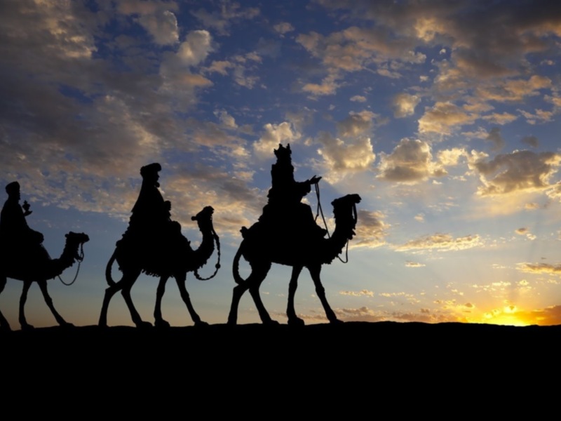 Tres jinetes en camello con sombreros, bajo un cielo dramático de nubes y puesta de sol.