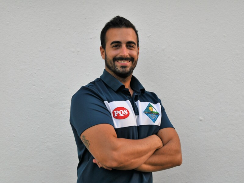 Un hombre con una camiseta azul y un logo de PQS en el brazo, posando con los brazos cruzados.