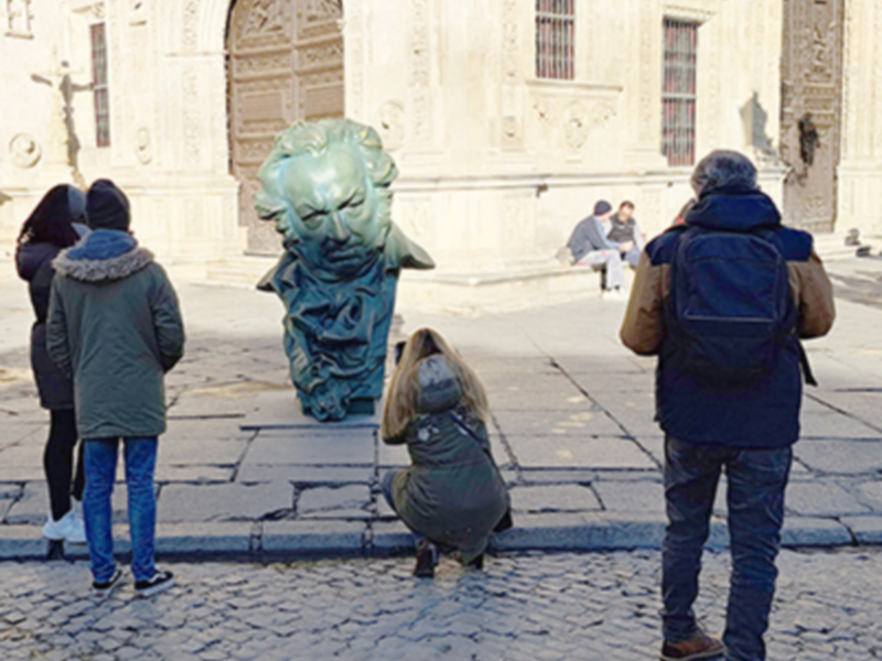 Turistas observando una escultura en el suelo frente a un edificio histórico.