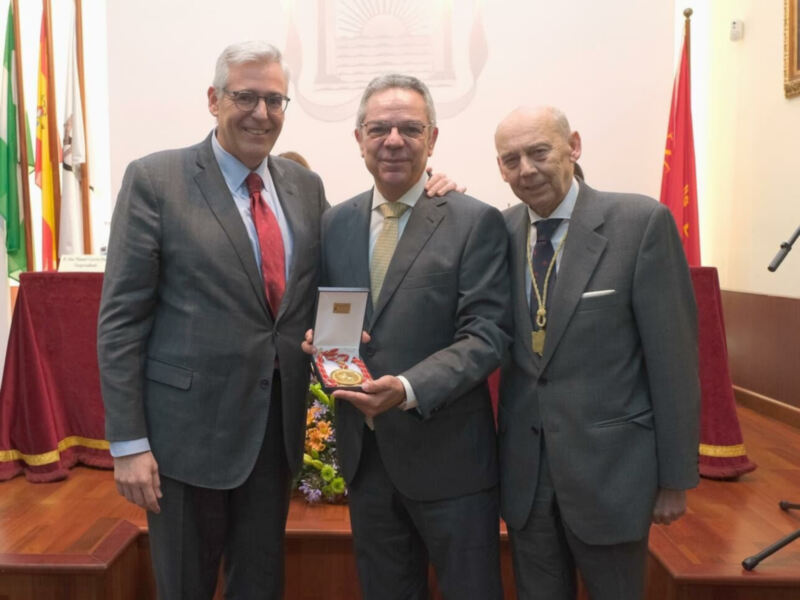 Tres hombres en trajes posan con una medalla en un evento formal en un salón con banderas.