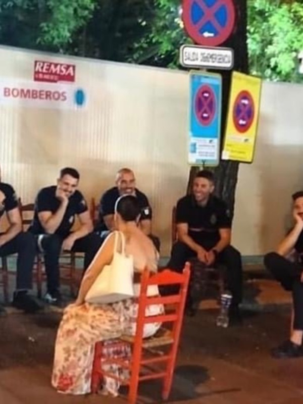 Un grupo de personas sentadas en sillas rojas frente a un edificio con carteles de señalización. La imagen muestra una escena casual, posiblemente en un parque o área pública.