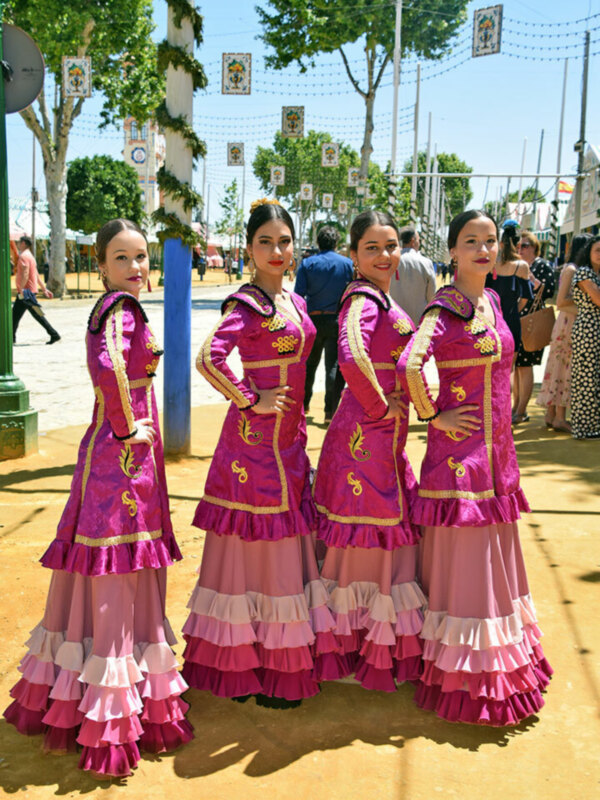 Tres mujeres vestidas con trajes tradicionales posan en una calle con árboles y banderas en el fondo.