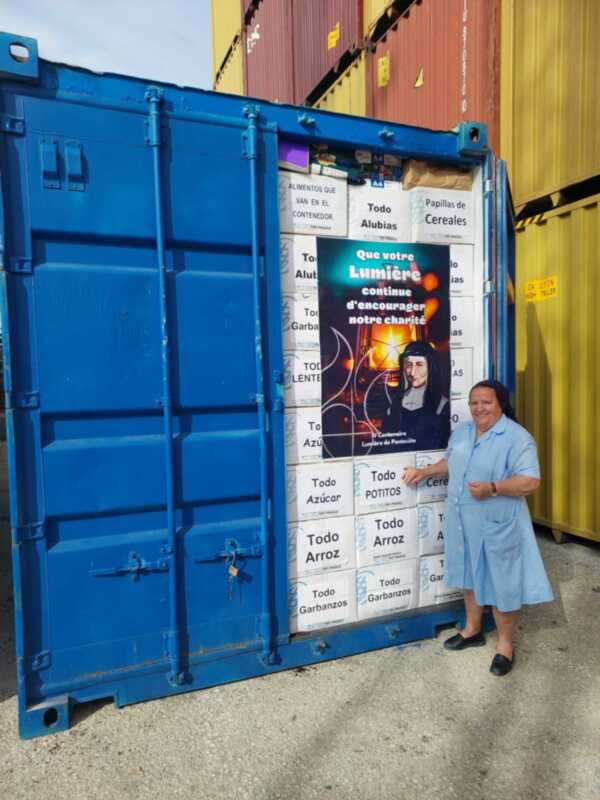 Una mujer frente a un contenedor azul lleno de cajas de alimentos, con un cartel en francés que dice 'Que votre Lumière continue d'encourager notre charité'.