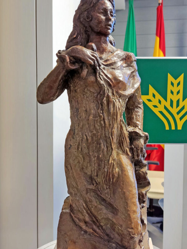 Sculpture of a woman in flowing attire, titled 'María de Trianas', displayed indoors with flags in the background.